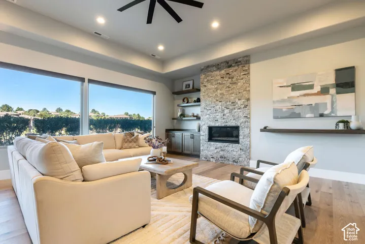 Tray ceiling featuring recessed lighting and a ceiling fan, stacked-stone fireplace, wood-finish flooring, and large windows with integrated shades