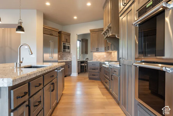 Chef's kitchen featuring custom wood cabinetry, granite countertops, stainless steel appliances including dual wall ovens, a large island with undermount sink and gooseneck faucet, pendant lighting, and warm wood-finish flooring throughout the open layout