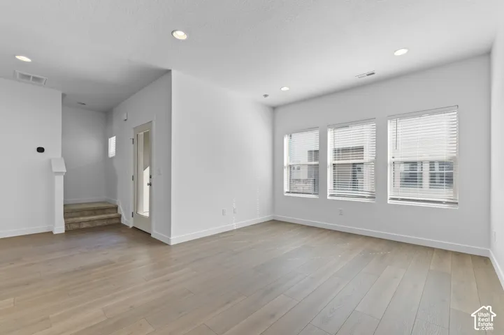 Open-concept interior featuring light wood-finish flooring, recessed lighting, triple-pane windows with horizontal blinds, and a glass-panel entry door