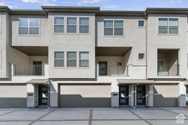 Multi-level exterior featuring light-toned stucco, white masonry accents, recessed balconies with horizontal railings, and multiple garage bays