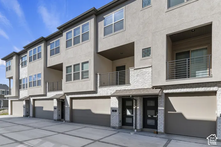 Contemporary multi-story exterior featuring a mix of stucco and stone-veneer siding, private recessed balconies with horizontal railings, multi-panel glass entry doors, and attached two-car garages