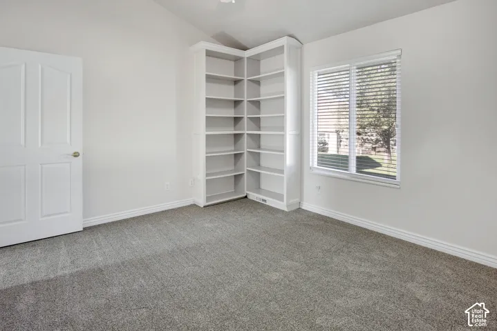 Vaulted ceiling, built-in corner shelving unit, neutral carpet flooring, and a window with horizontal blinds