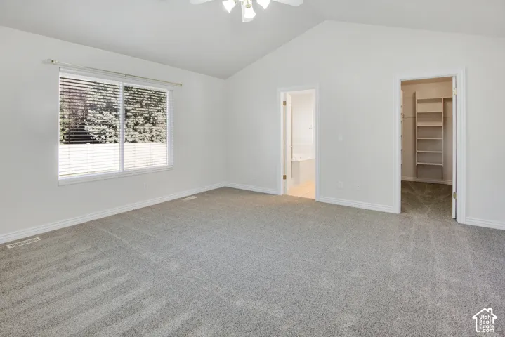 Vaulted ceilings, neutral carpeting, large window with horizontal blinds, ceiling fan, and walk-in closet featuring built-in shelving