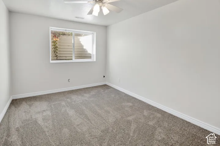 Neutral-toned interior space featuring light-colored wall paint, wall-to-wall carpeting, an integrated ceiling fan with light fixture, and a sliding window with an external egress window well