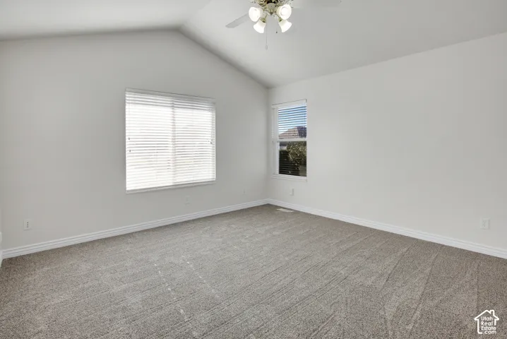 Vaulted ceilings, neutral carpeting, and multi-light ceiling fan fixture, complemented by dual windows with horizontal blinds