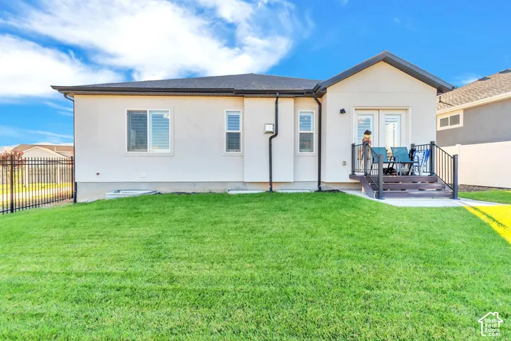 Rear exterior featuring stucco siding, gabled roofline, manicured lawn, metal privacy fencing, and elevated composite stairs leading to double glass doors