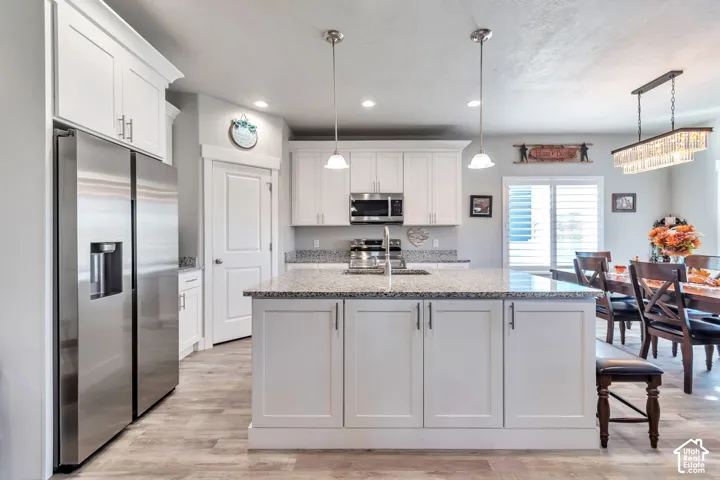 Kitchen featuring white shaker-style cabinetry, stone countertops, a central island with an integrated sink, stainless steel appliances, and wood-finish flooring