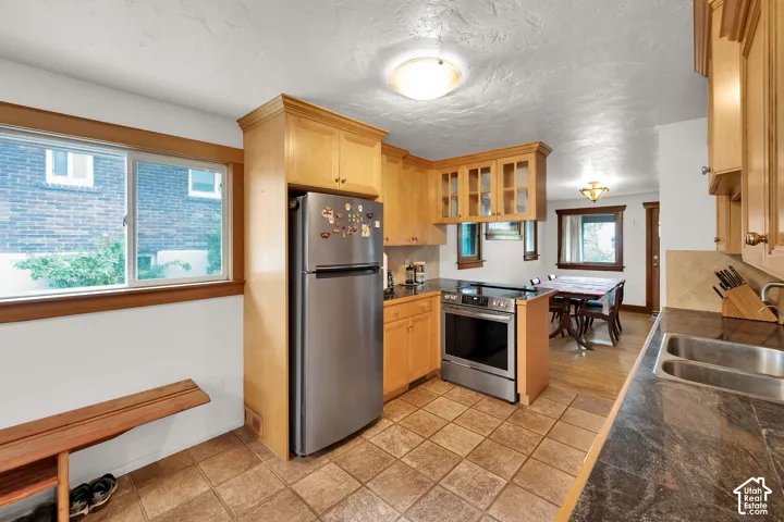 Kitchen featuring light wood cabinetry with glass-panel display uppers, stainless steel appliances, dark stone-finish countertops, dual-basin stainless steel sink, and ceramic tile flooring