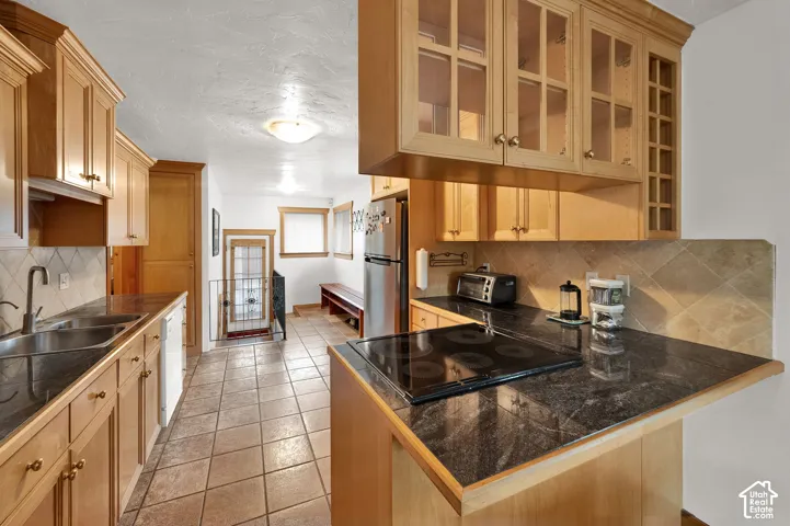 Kitchen featuring natural wood cabinetry with glass-front uppers, dark stone countertops, diamond-pattern tile backsplash, and tile flooring