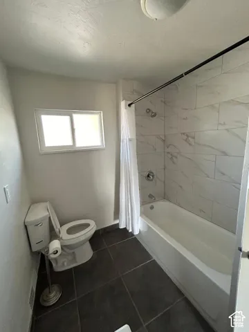 Full bathroom featuring large-format dark tile flooring, a bathtub-shower combination with floor-to-ceiling white tiled surround, and a white porcelain toilet
