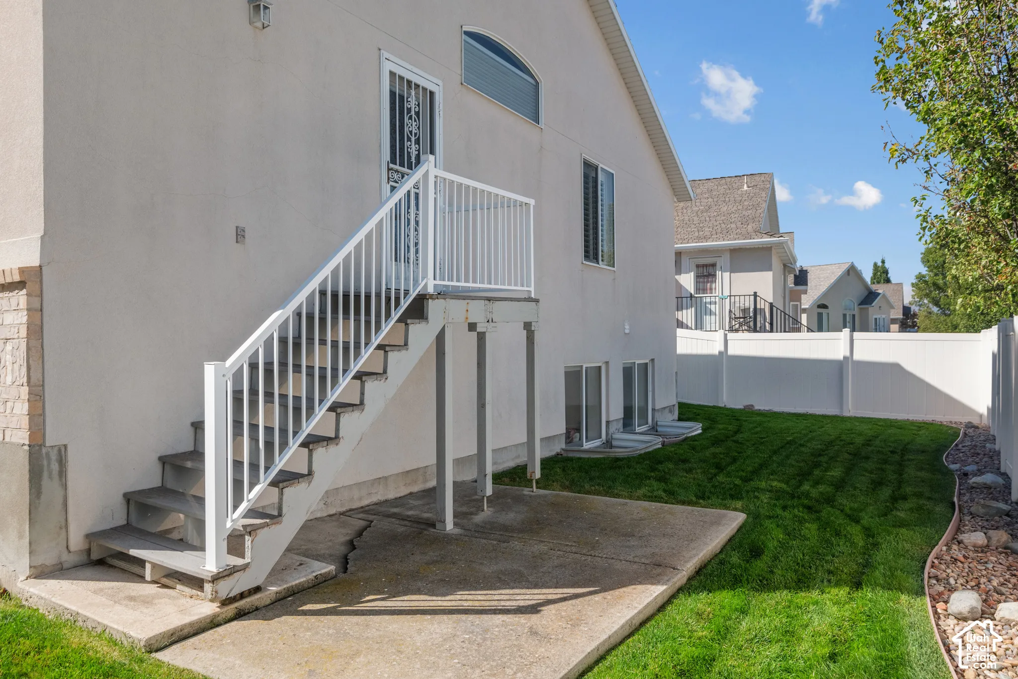 Exterior stucco facade with elevated deck, white metal railing, concrete patio, and fenced backyard featuring a manicured lawn