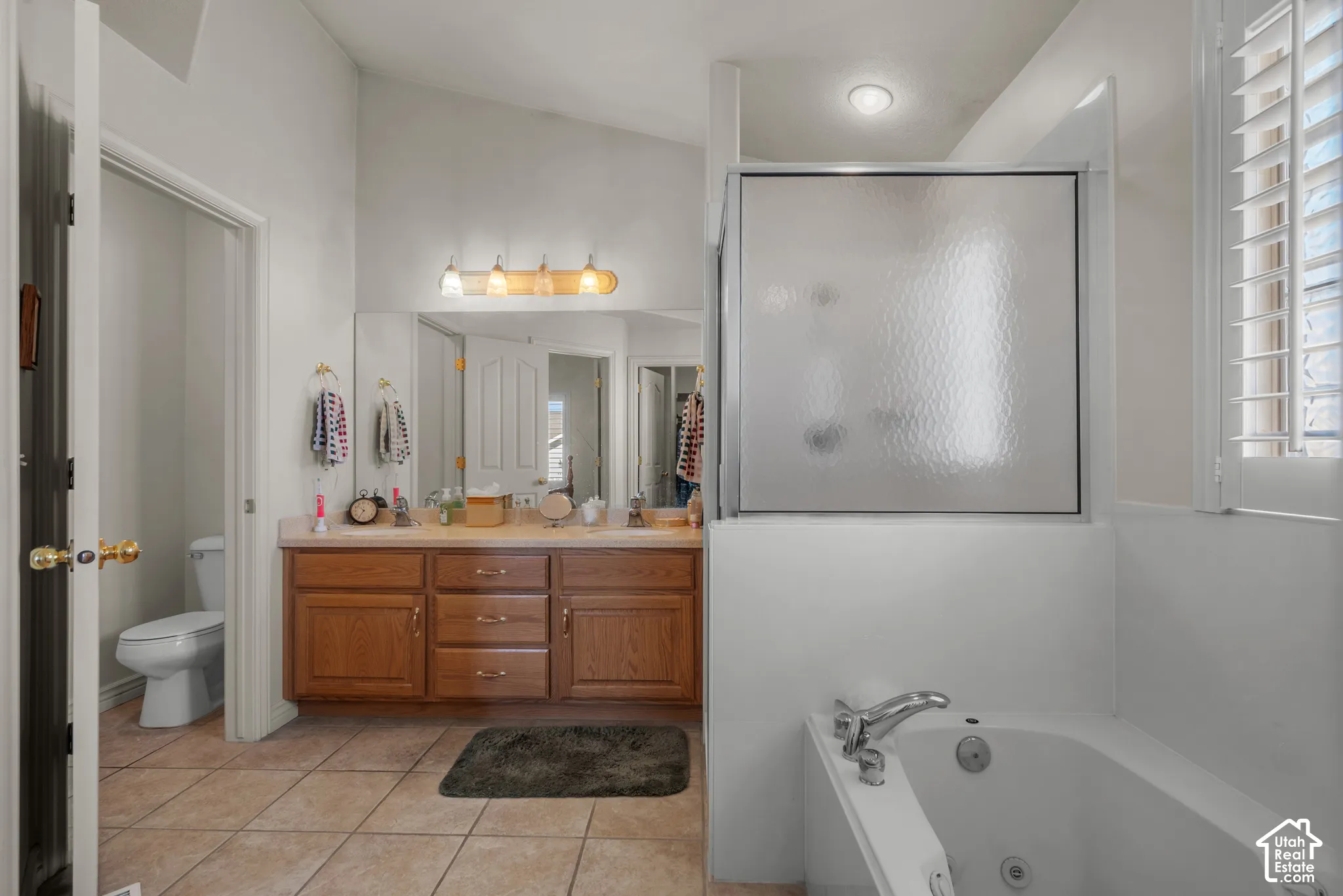 Bathroom featuring dual-sink vanity with wood cabinetry, tile flooring, built-in jetted tub, and a separate glass-enclosed shower