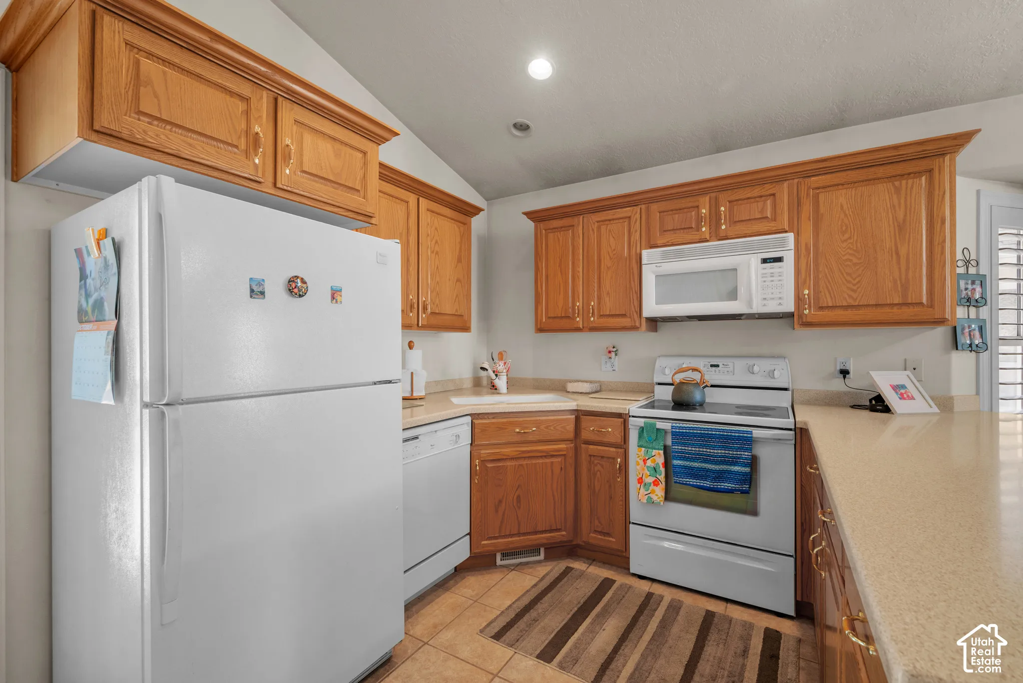 Kitchen featuring oak cabinetry, light-toned solid surface countertops, a white built-in dishwasher, an over-the-range microwave, and ceramic tile flooring