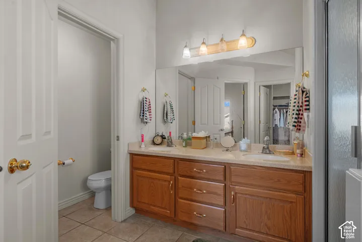 Dual-sink vanity with wood-finish cabinetry, solid-surface countertop, full-width mirror, four-light wall fixture, and tile flooring