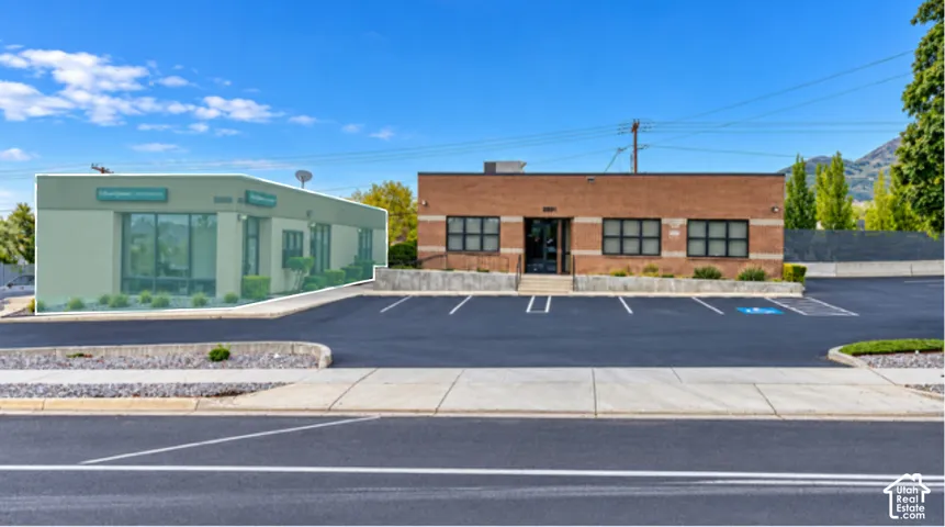 Commercial property exterior featuring a brick facade, multi-pane windows, paved asphalt parking lot, stone retaining wall, and mountain backdrop