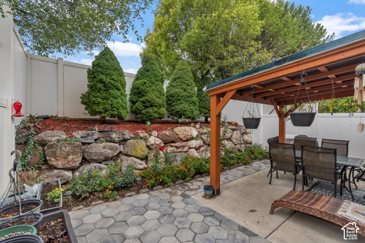 Outdoor patio featuring a hexagonal paver hardscape, a covered timber-framed pergola, and a tiered stone retaining wall with mature evergreen landscaping and mulch beds