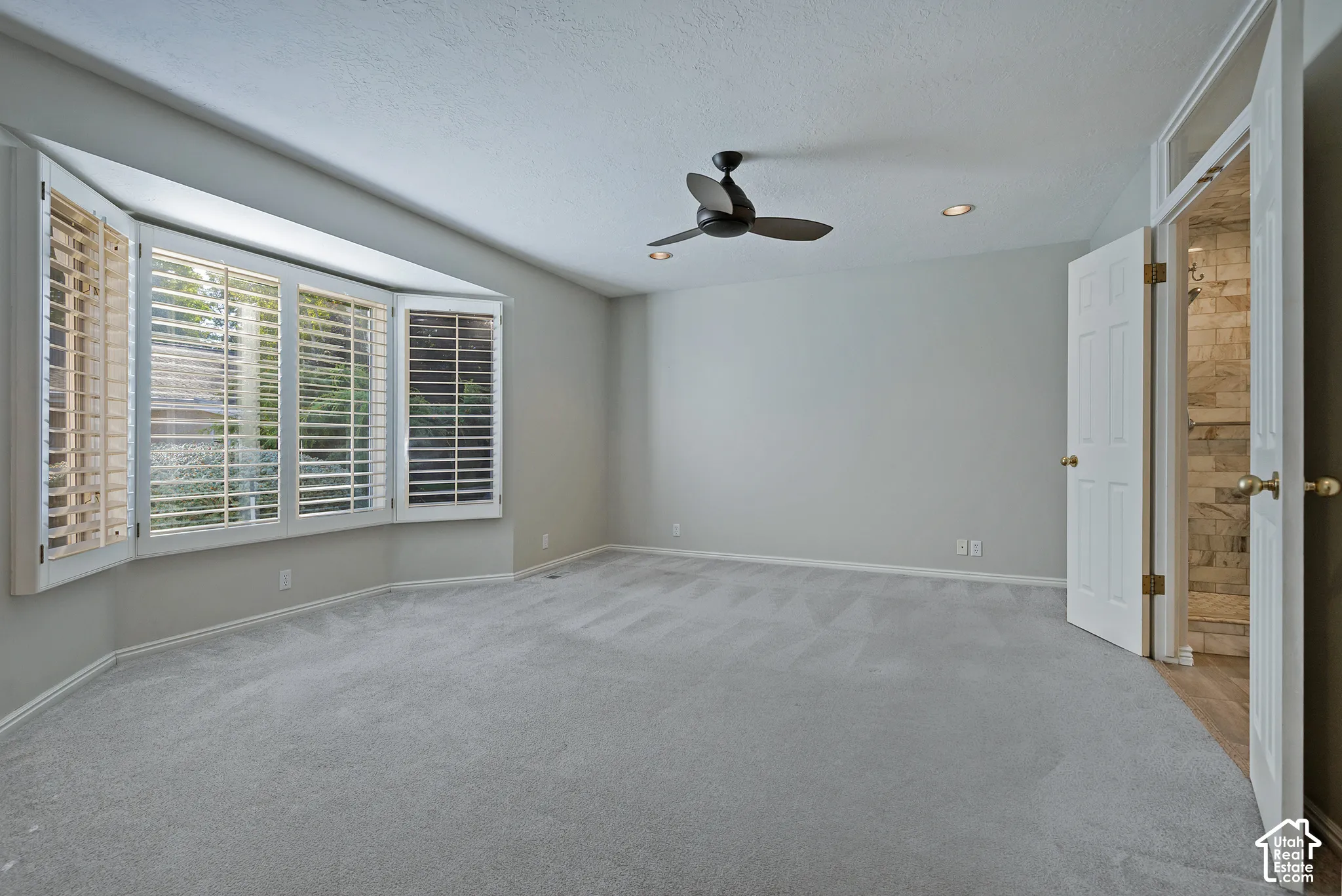 Bay window with plantation shutters, ceiling fan, recessed lighting, carpeted flooring, and an adjoining room with stone-finish walls