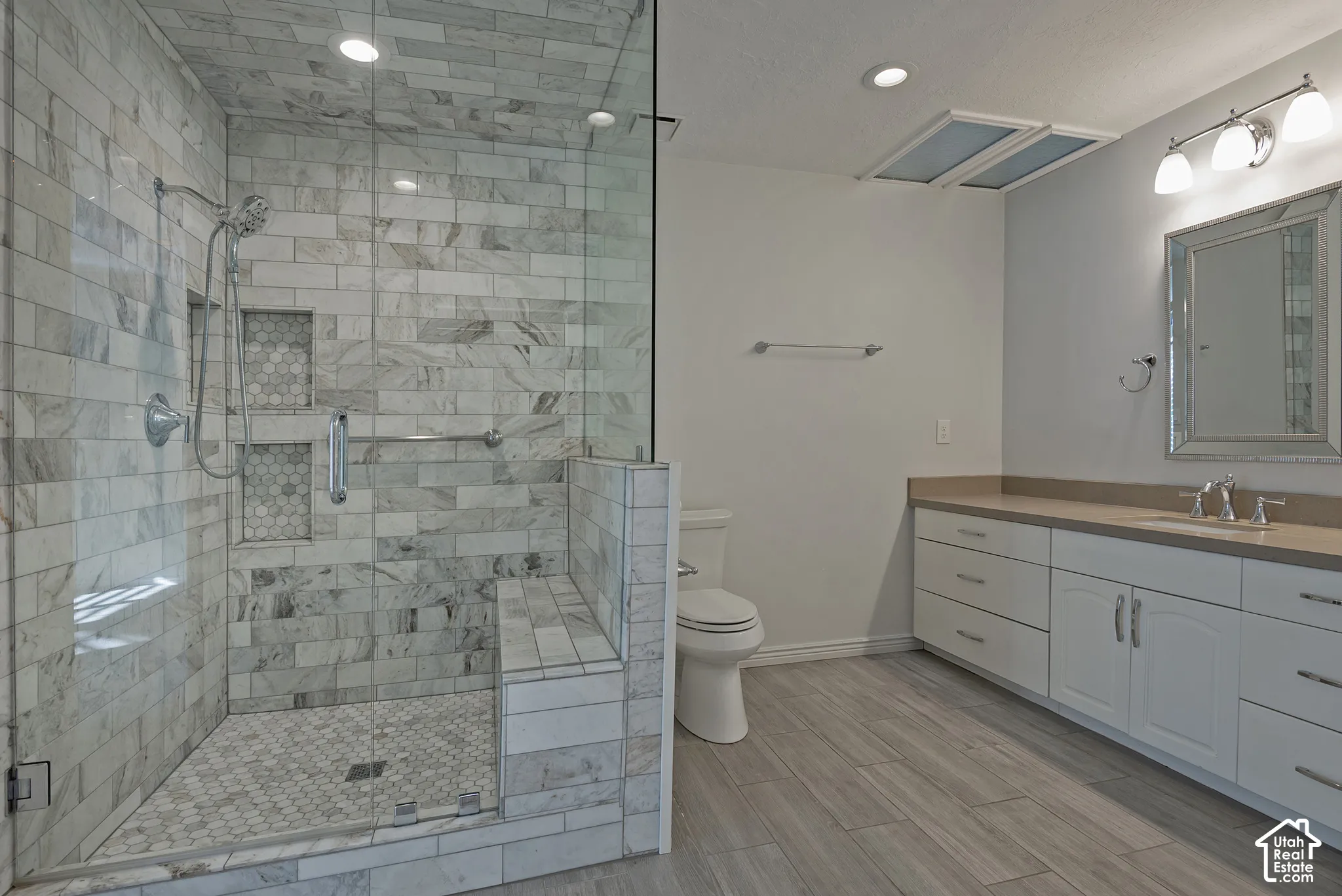Glass-enclosed shower with subway tile and built-in bench, dual recessed shower niches, wood-finish flooring, an extended vanity with dual sinks, and white cabinetry