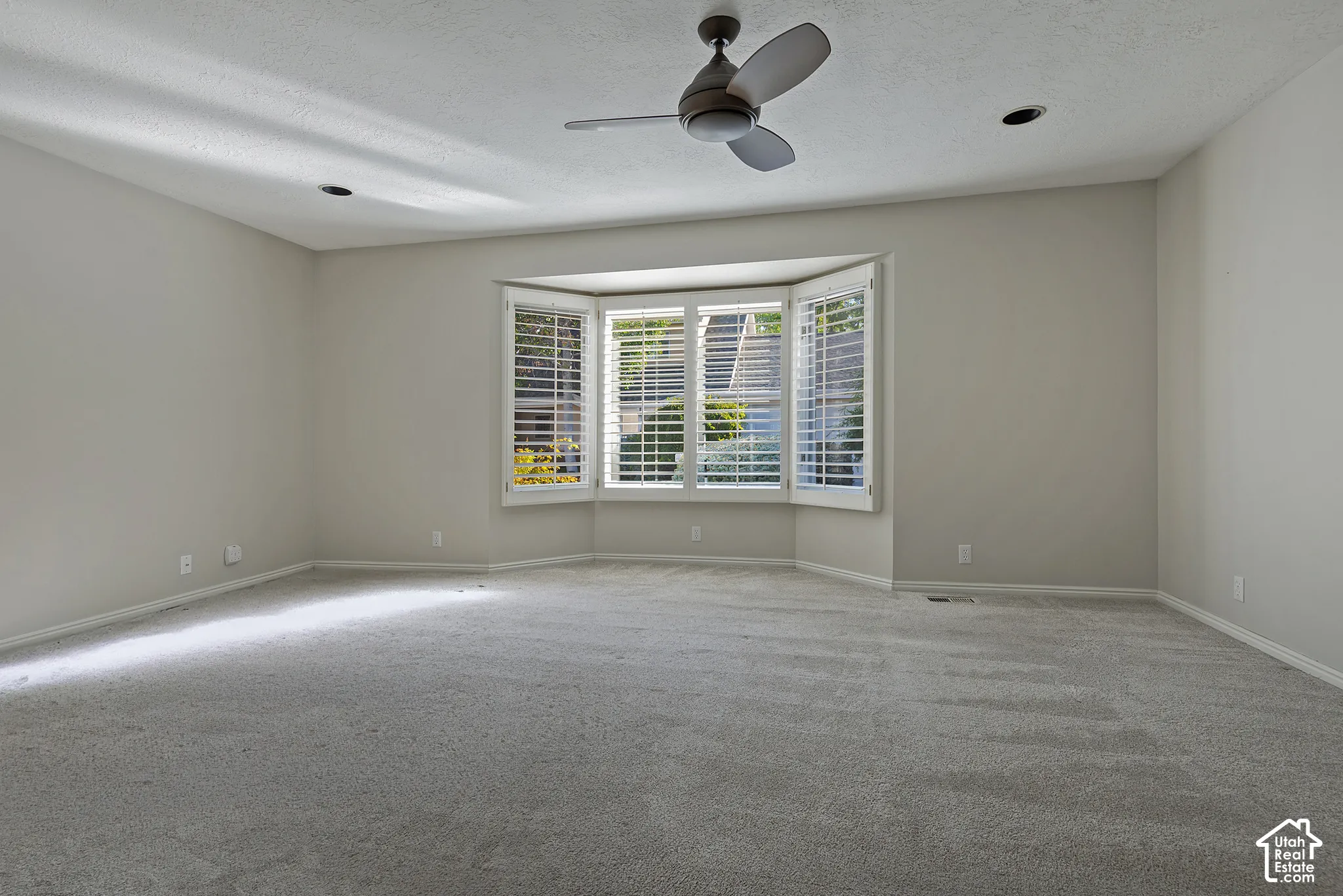 Spacious room featuring a bay window with white plantation shutters, neutral carpet, recessed lighting, and a ceiling fan