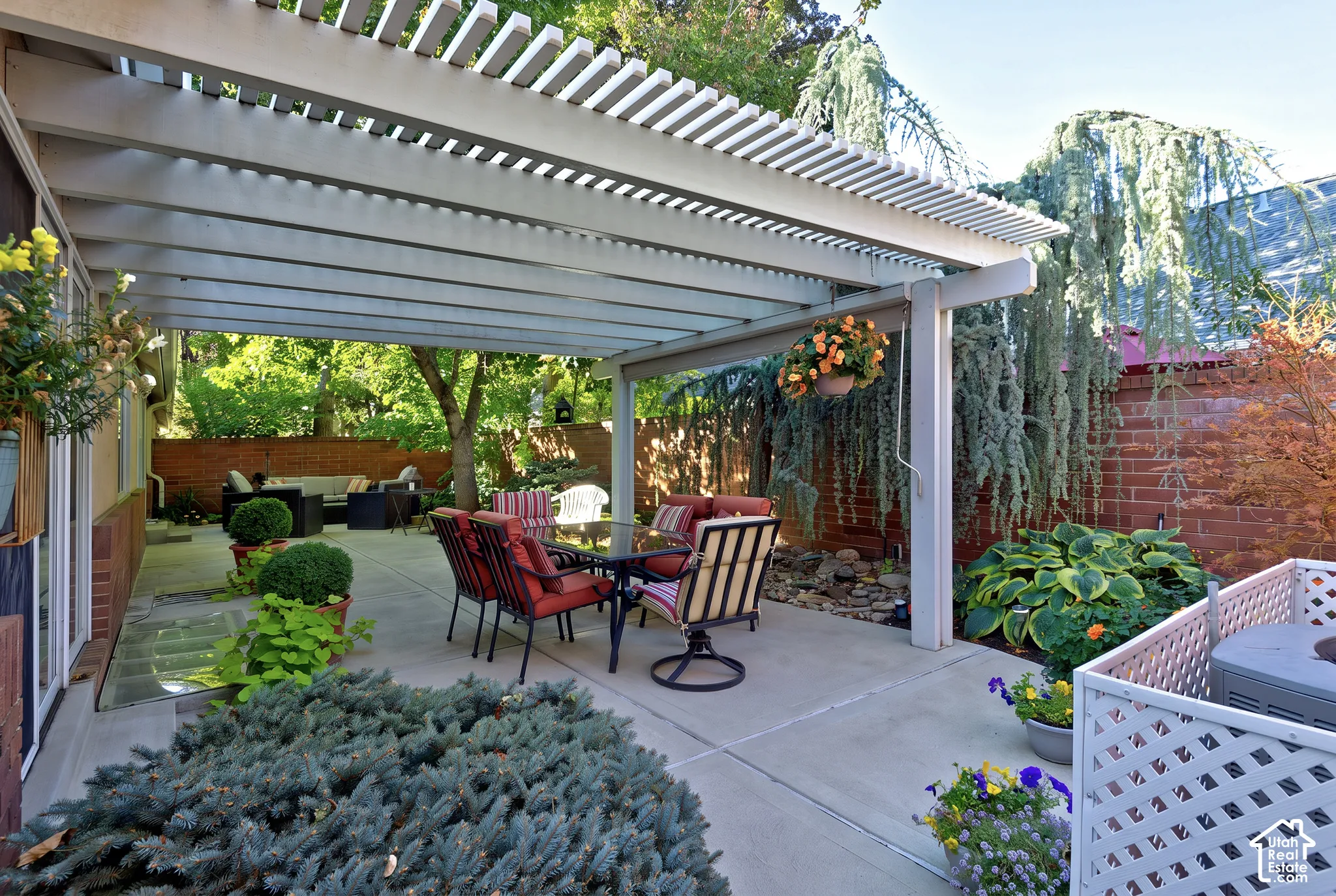 Concrete patio featuring a slatted pergola, brick wall, and mature landscaping
