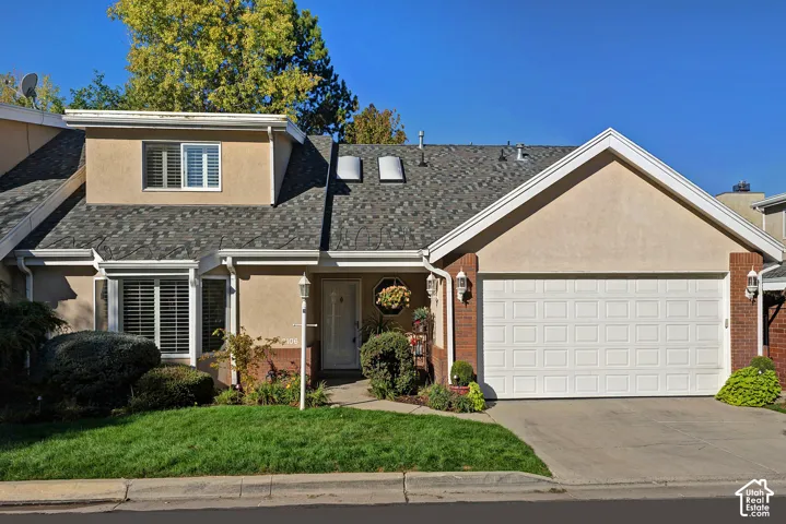 Stucco exterior with brick accents, attached garage with panel door, asphalt shingle roof with skylights, dormer window with louvered shutters, and front entryway with a decorative light fixture