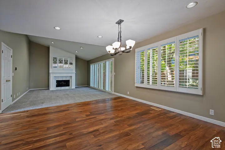 Wood-finish flooring leading to carpeted areas, a fireplace with a mantel and integrated cabinetry, a multi-pane window with plantation shutters, recessed lighting, and a chandelier