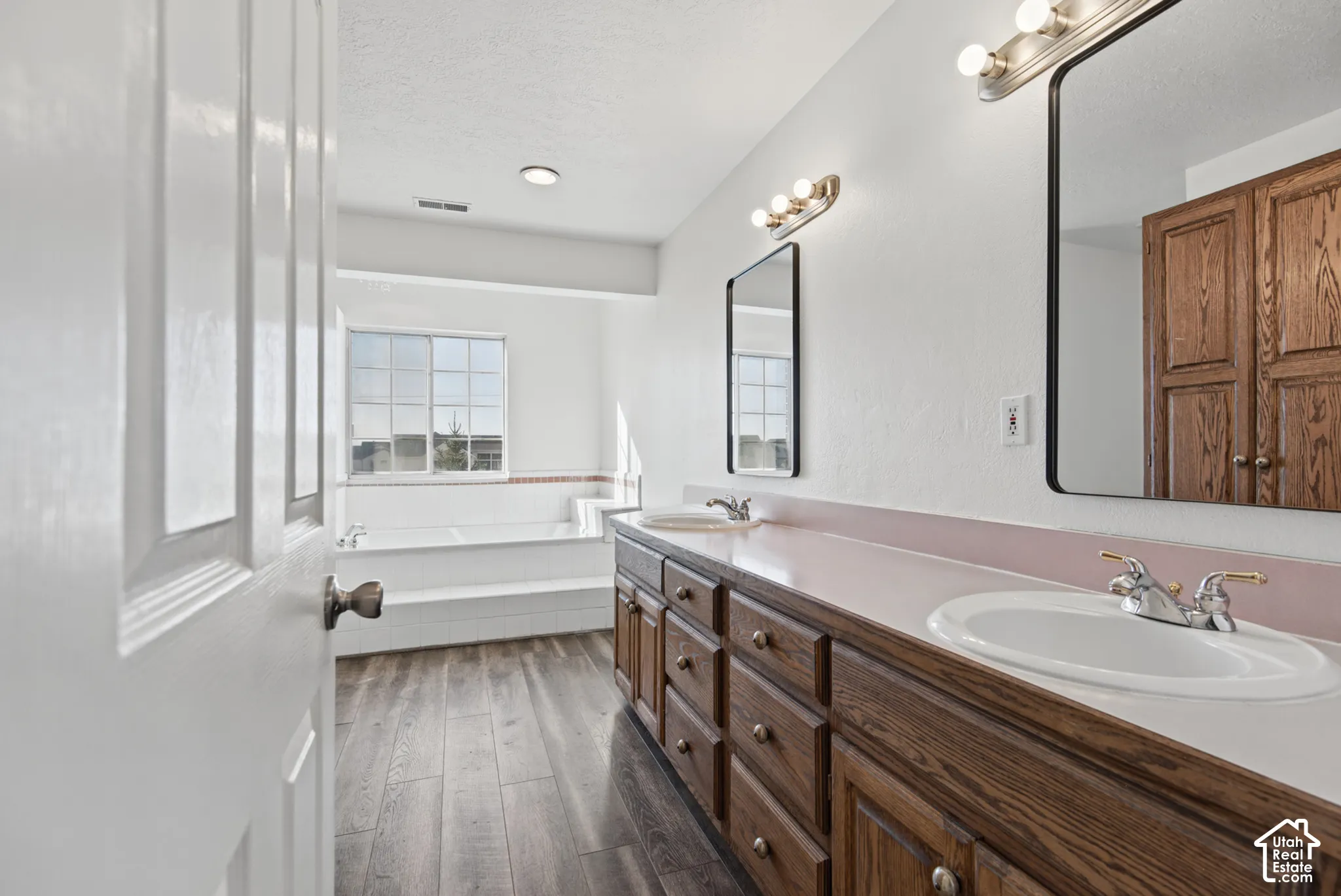 Dual-sink vanity with wood-finish cabinetry, countertop basins, and chrome hardware, paired with wood-finish flooring and a built-in soaking tub