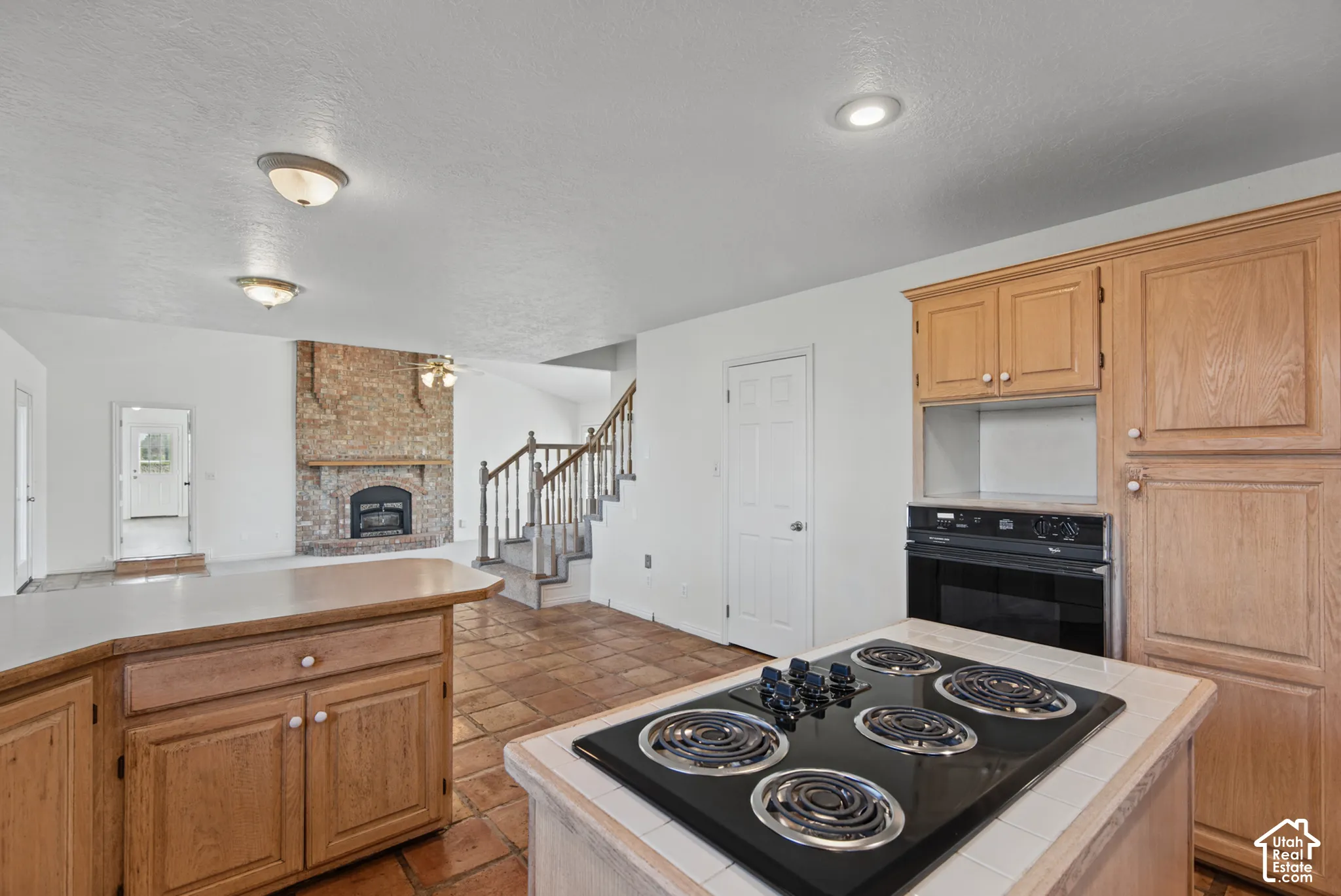 Kitchen featuring wood cabinetry, a tiled island countertop with a built-in cooktop, tile flooring, a brick fireplace, and a wooden staircase