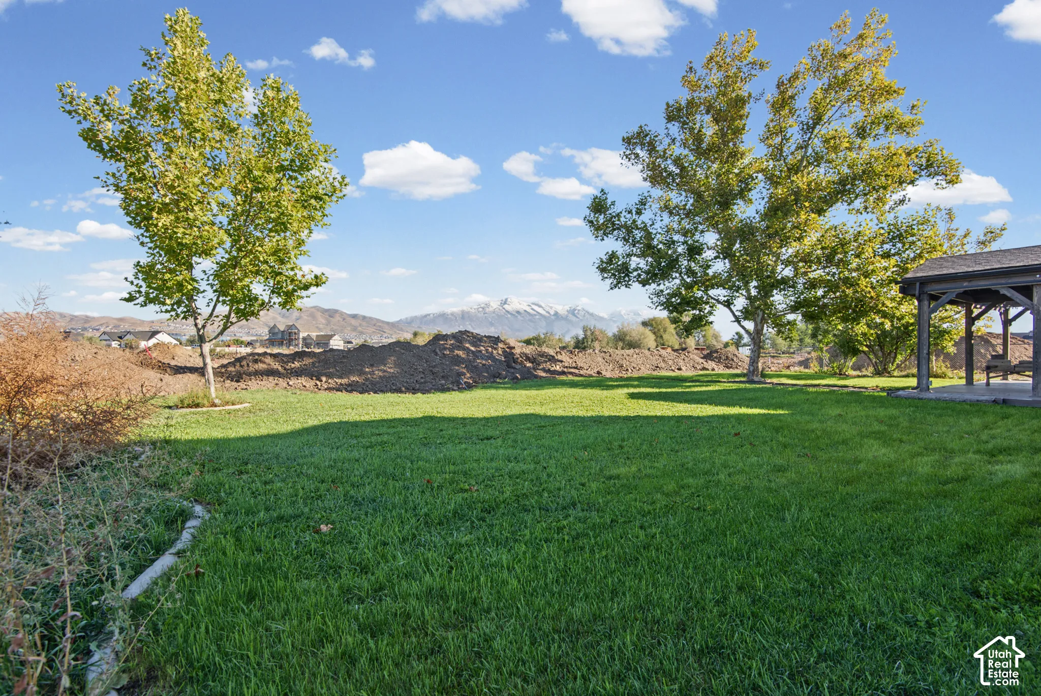 Expansive backyard featuring a manicured lawn, mature trees, a covered patio structure, and a mountain backdrop