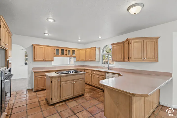 Kitchen featuring natural wood cabinetry, a central island with a built-in cooktop, tile flooring, recessed lighting, and an arched window