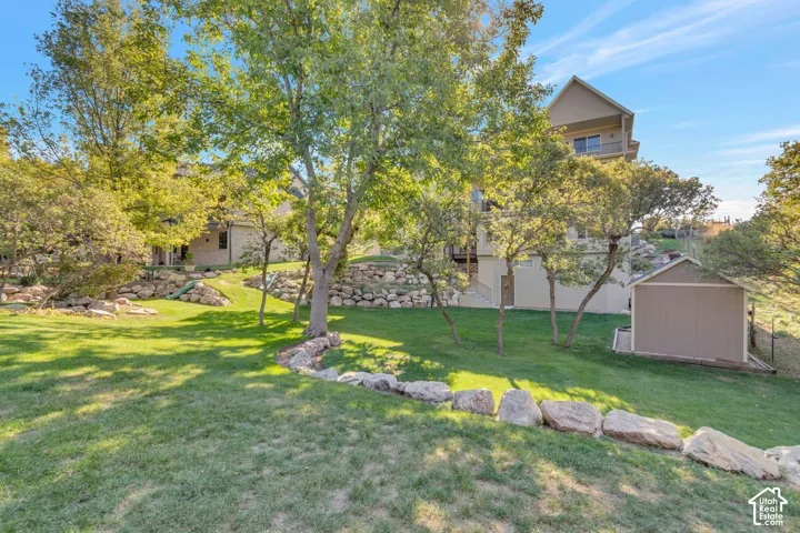 Terraced backyard featuring natural boulder retaining walls, expansive lawn, mature trees, and an exterior storage structure