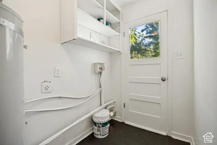 Service area featuring dark flooring, wall-mounted open shelving, a water heater, and a white exterior door with a glass pane
