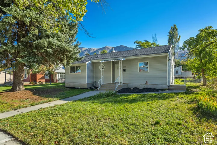 Single-story residence featuring light-toned horizontal siding, an asphalt shingle roof, a covered front entryway with decorative metal railings, and a concrete walkway set against a mountain backdrop