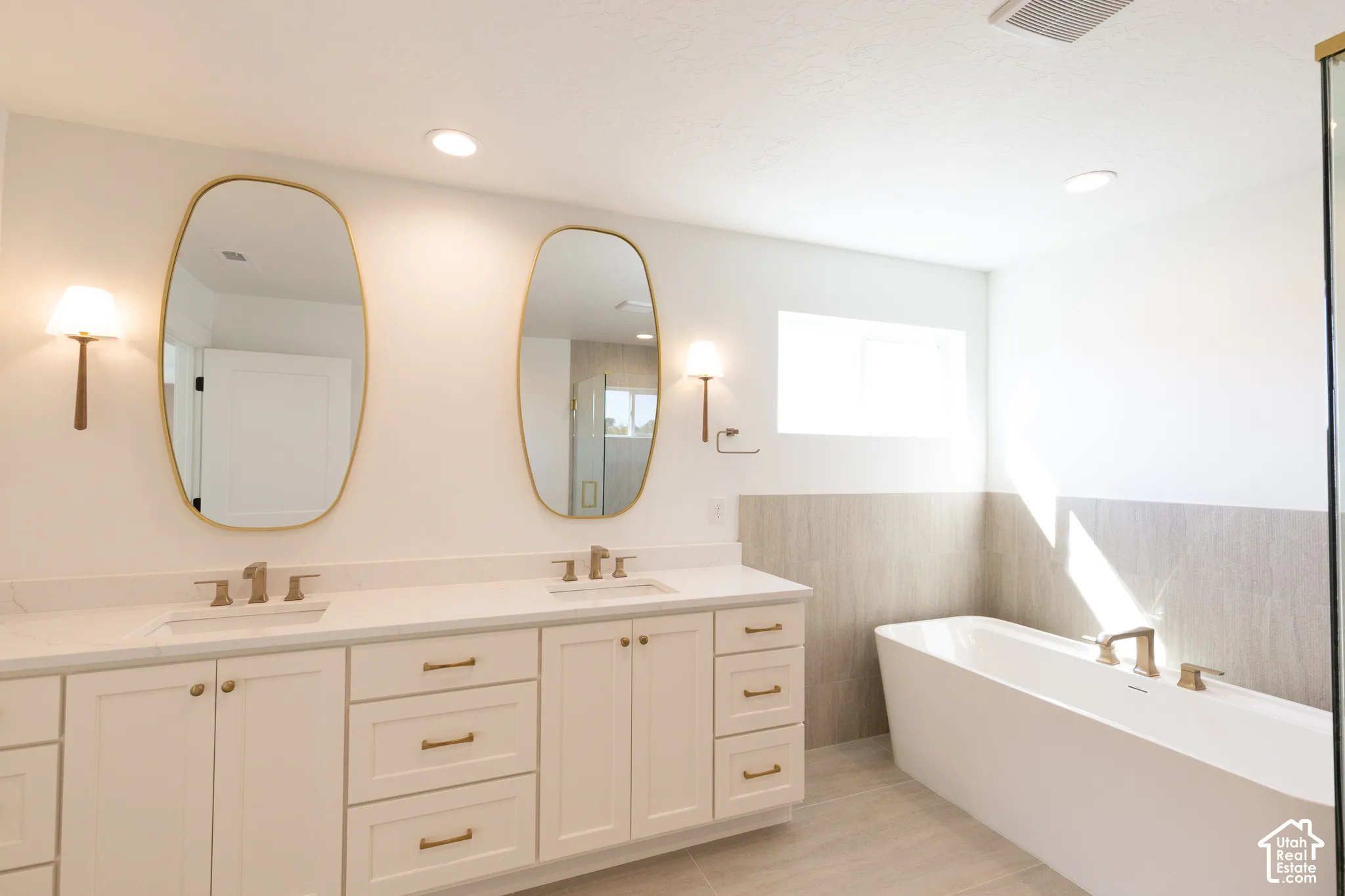 Dual-sink vanity with light-toned cabinetry, solid surface countertop, metallic hardware, oval wall-mounted mirrors, and a freestanding soaking tub with tile surround