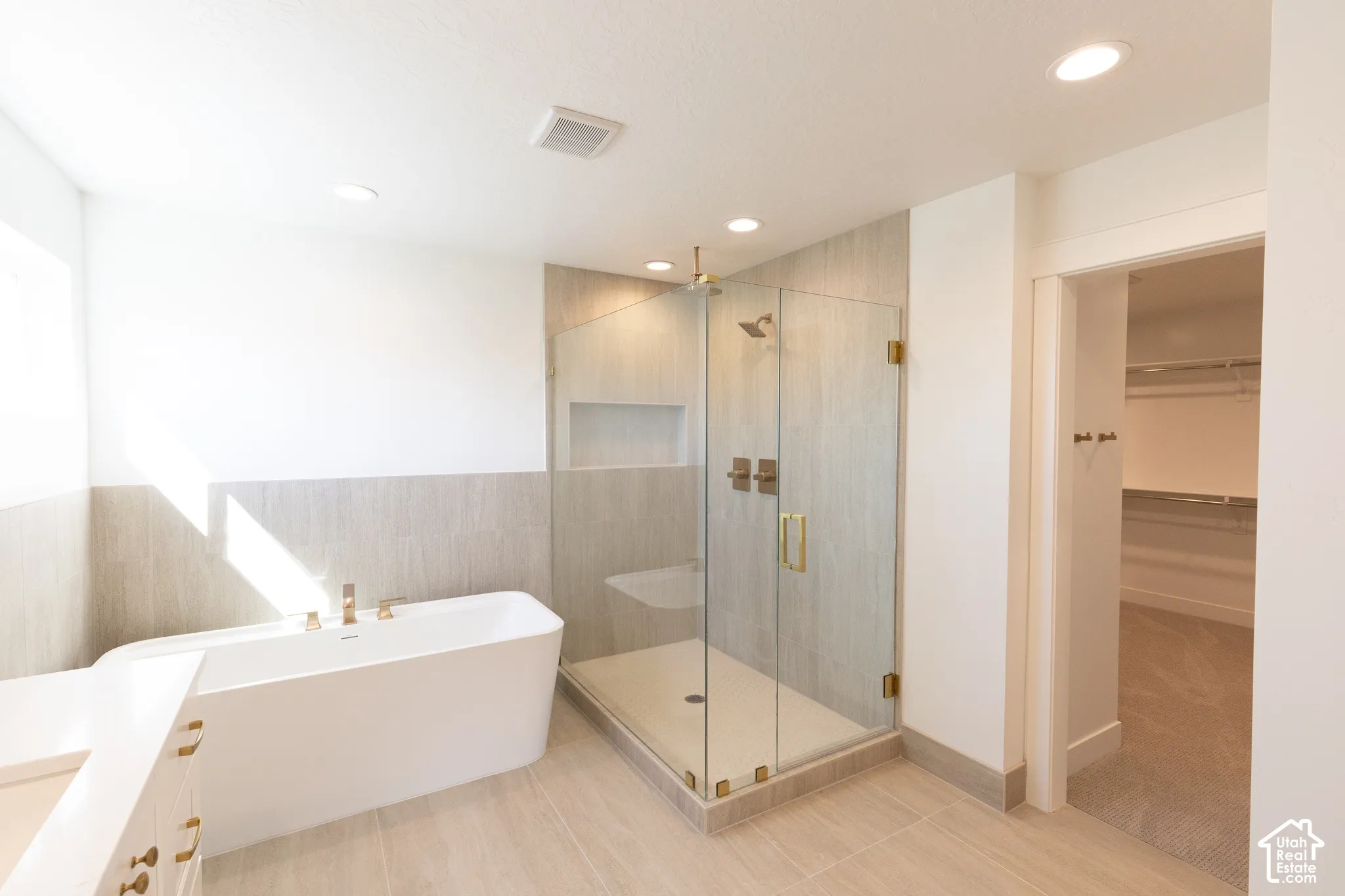 Freestanding soaking tub with polished metal fixtures, glass-enclosed shower featuring tile surrounds and a built-in recessed shelf, recessed lighting, and large-format tile flooring