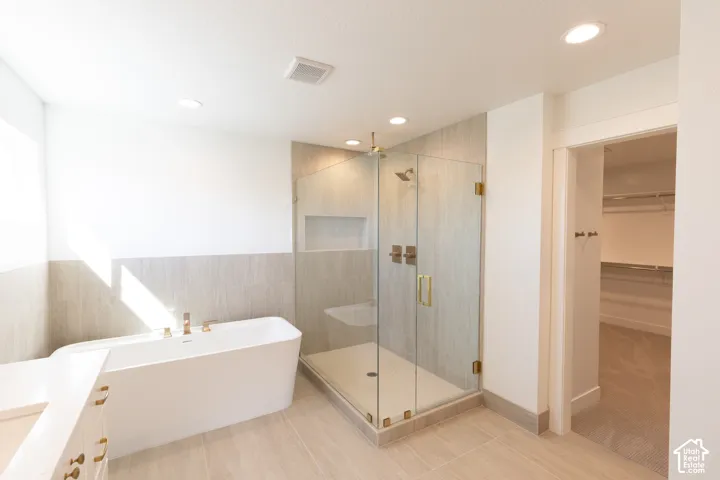 Freestanding soaking tub with polished metal fixtures, glass-enclosed shower featuring tile surrounds and a built-in recessed shelf, recessed lighting, and large-format tile flooring