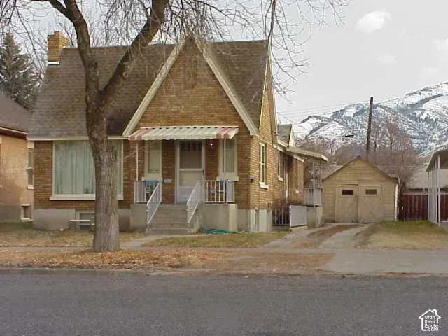 Brick exterior featuring a steep-pitched gable roof, front porch with metal awning, concrete walkway, detached two-car garage, and mountain backdrop