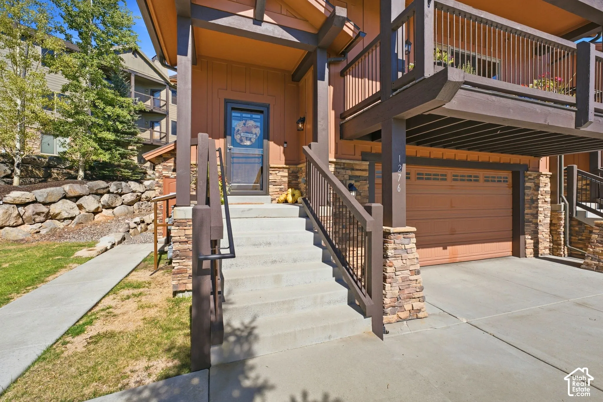 Multi-level exterior featuring stone veneer accents, wood-finish siding, a private balcony with metal railings, a concrete stairway, and an attached garage