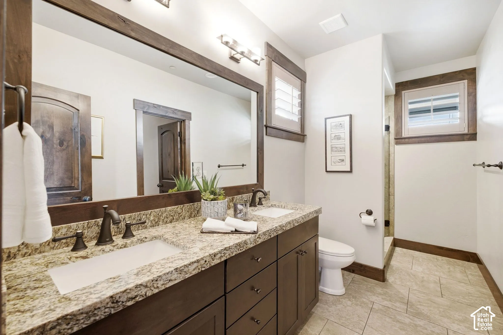 Dual-sink vanity with stone countertops, dark wood cabinetry, tile flooring, and matching wood-trimmed mirrors