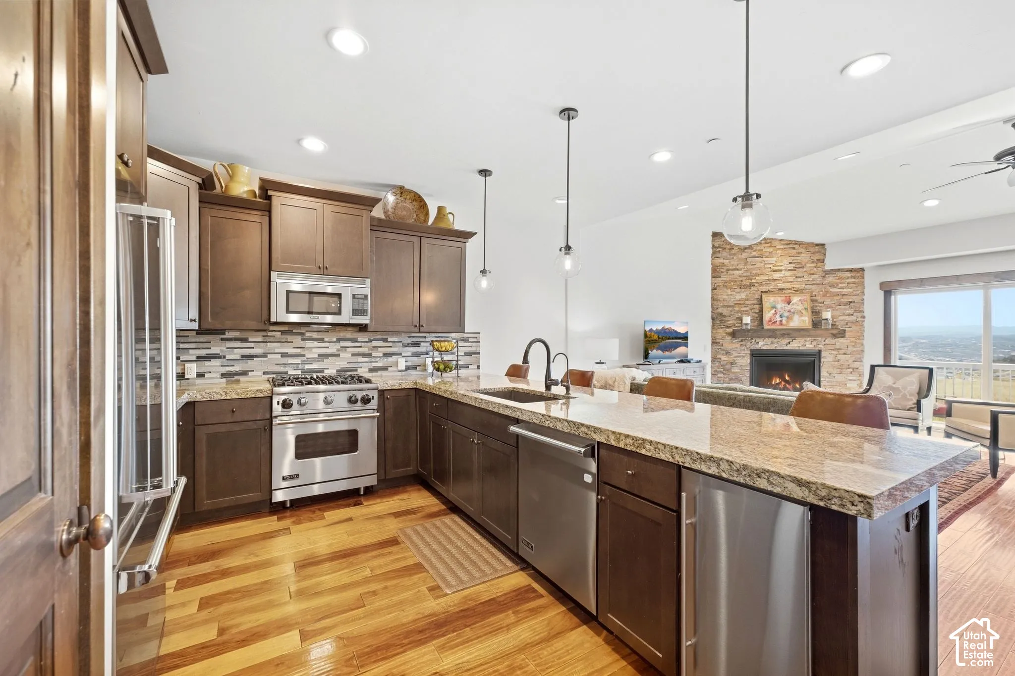 Kitchen featuring dark wood cabinetry, stone-finish countertops, stainless steel appliances, stacked-stone fireplace, and wood-finish flooring
