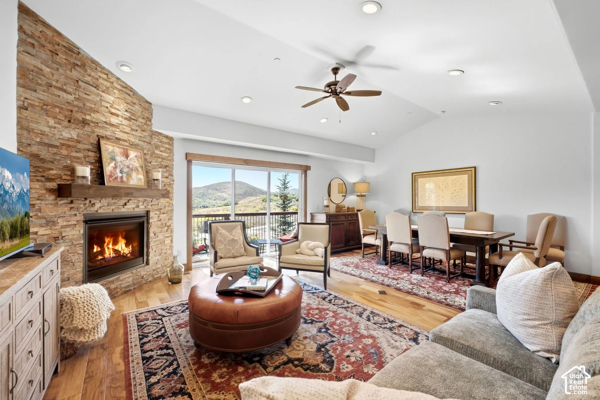 Floor-to-ceiling stone fireplace with timber mantel, wood-finish flooring, vaulted ceiling with recessed lighting, and sliding glass doors opening to an exterior deck
