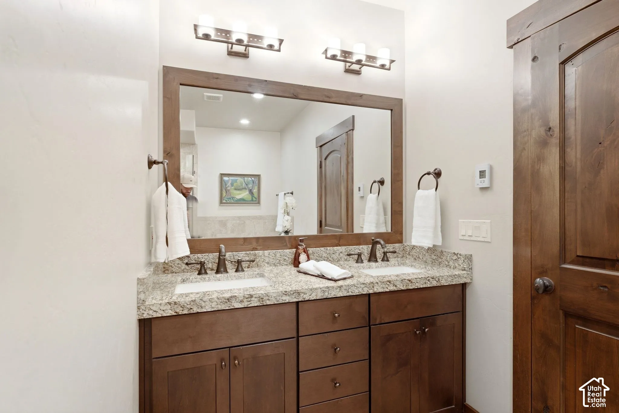 Double vanity featuring stained wood cabinetry, stone-finish countertop with dual undermount sinks, bronze-tone fixtures, and a framed mirror with integrated wall lighting