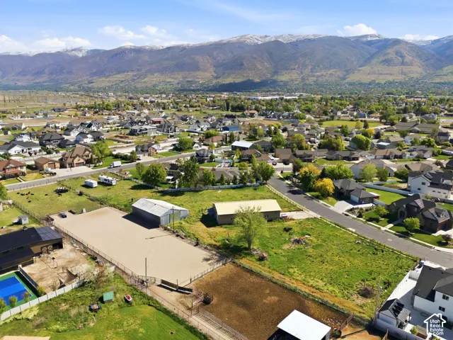 Aerial perspective featuring fenced equestrian grounds, two metal outbuildings, a blue-surface sport court, and prominent mountain backdrop