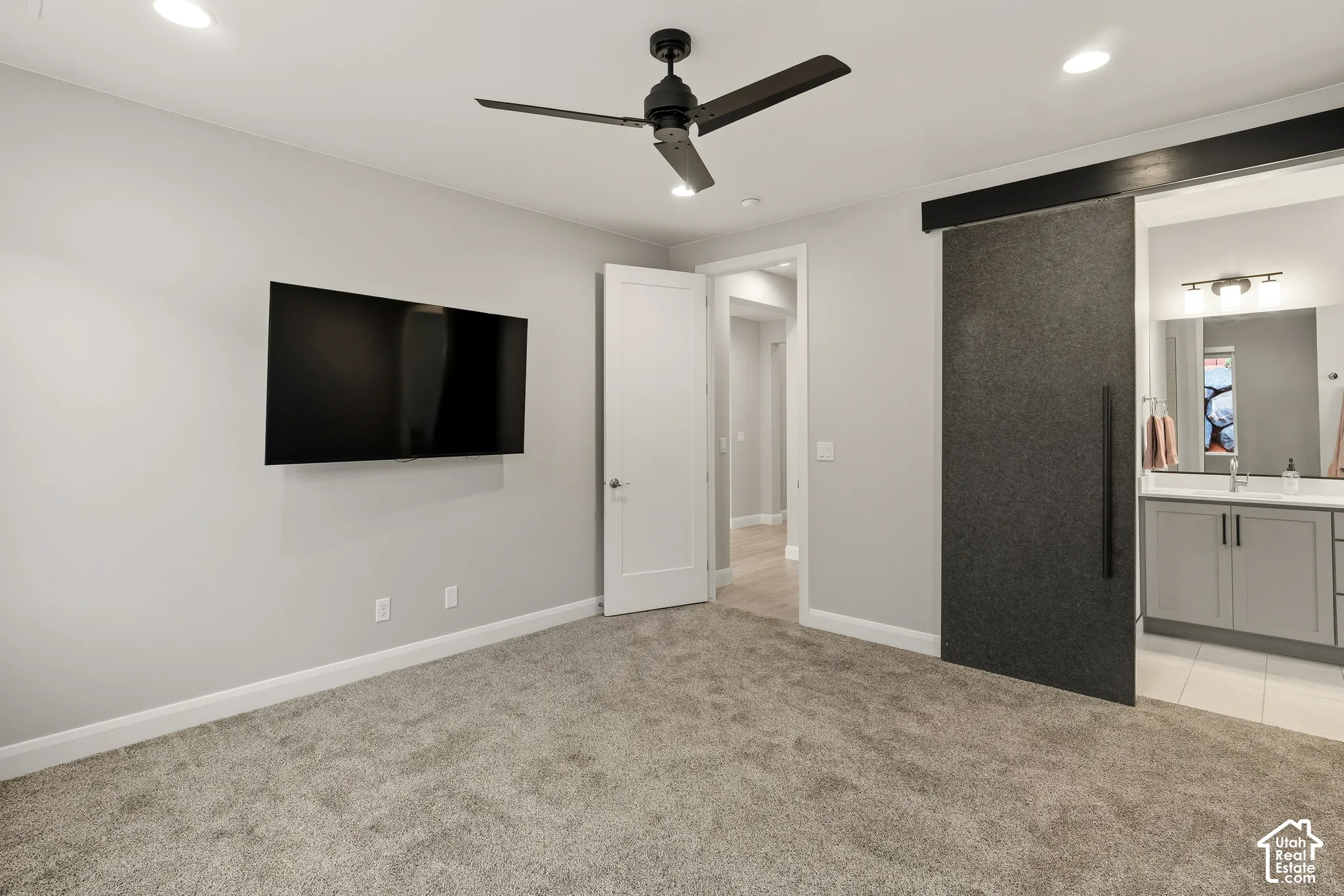 Room interior featuring neutral wall paint, charcoal-colored sliding barn door, recessed lighting, matte black ceiling fan, and grey cabinetry with a rectangular mirror