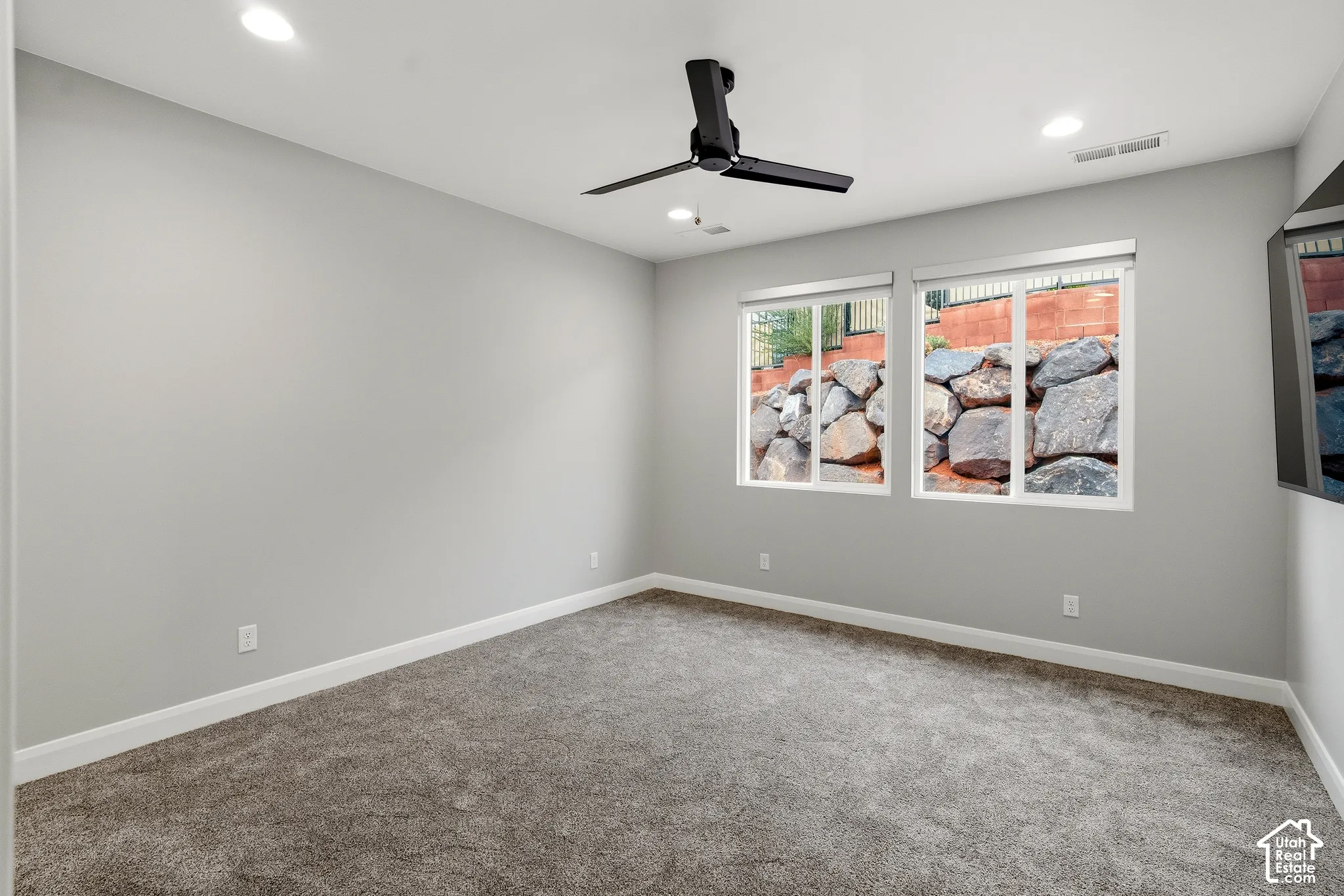Neutral-toned interior featuring wall-to-wall carpeting, recessed lighting, a matte black ceiling fan, and dual windows showcasing a stone retaining wall