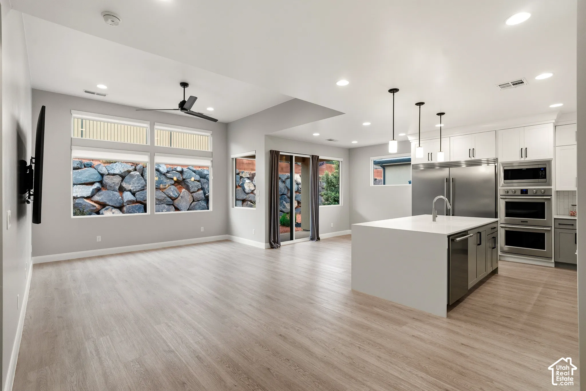 Open-concept interior featuring light wood-finish flooring, a center island with solid surface countertop, stainless steel double wall ovens, recessed lighting, and large windows with integrated rock wall exterior elements