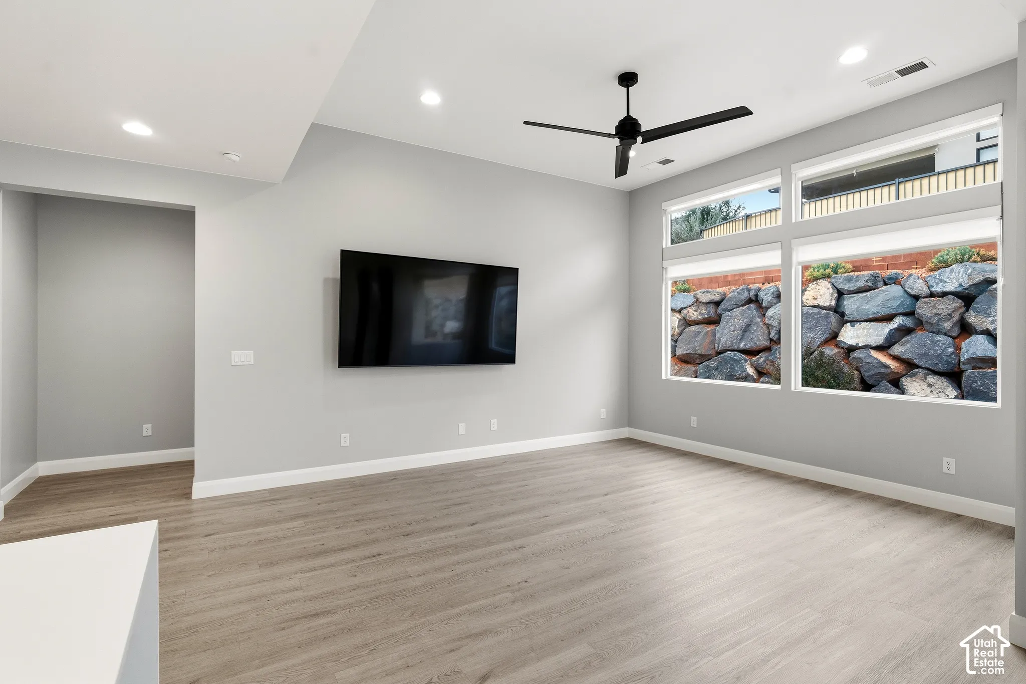 Living area featuring wood-finish flooring, recessed lighting, matte black ceiling fan, and large windows with exposure to a tiered stone retaining wall