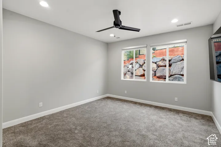 Neutral-toned interior featuring wall-to-wall carpeting, recessed lighting, a matte black ceiling fan, and dual windows showcasing a stone retaining wall