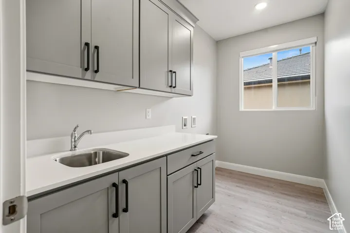 Utility area featuring shaker-style grey cabinetry, solid-surface countertops, an undermount stainless steel sink with a chrome faucet, and wood-finish flooring