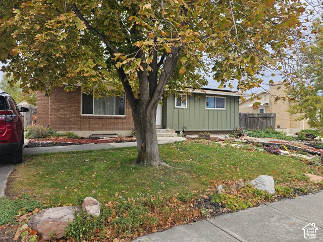 Exterior facade featuring a combination of brick and vertical siding, mature trees, and landscaping elements including rock features and shrubbery